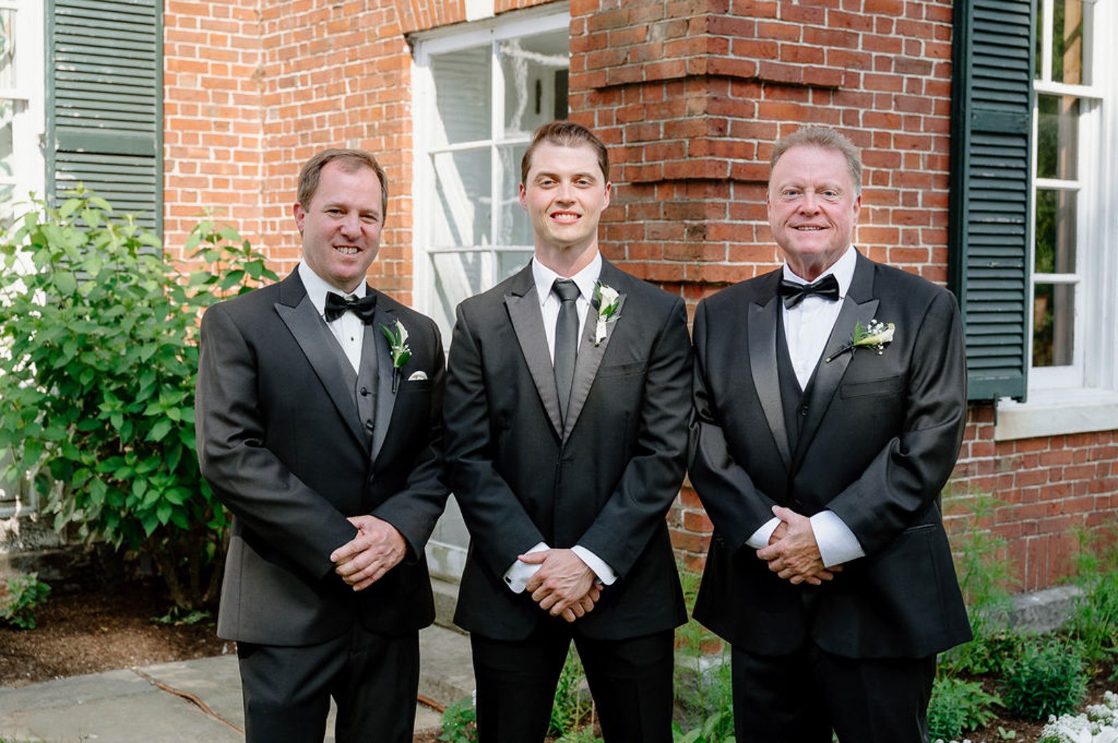 The groom, his father, and his bride's father in front of a brick house. 