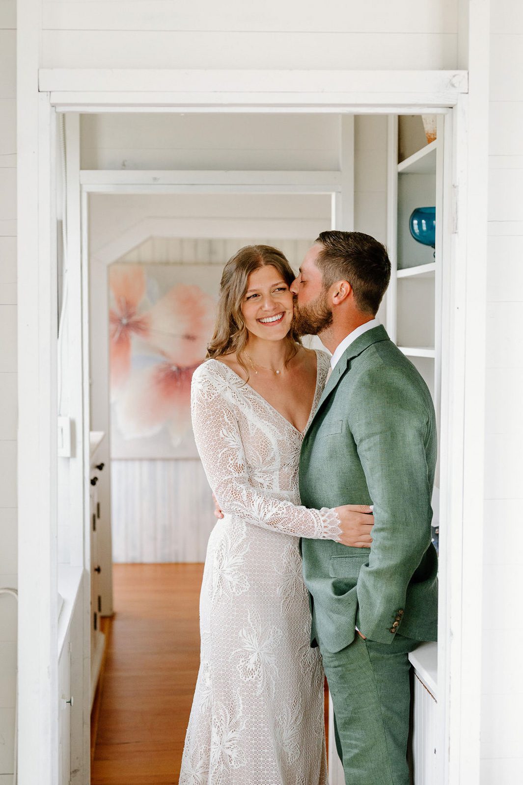 the groom kissing is wife on the cheek as she smiles, both in a doorway.