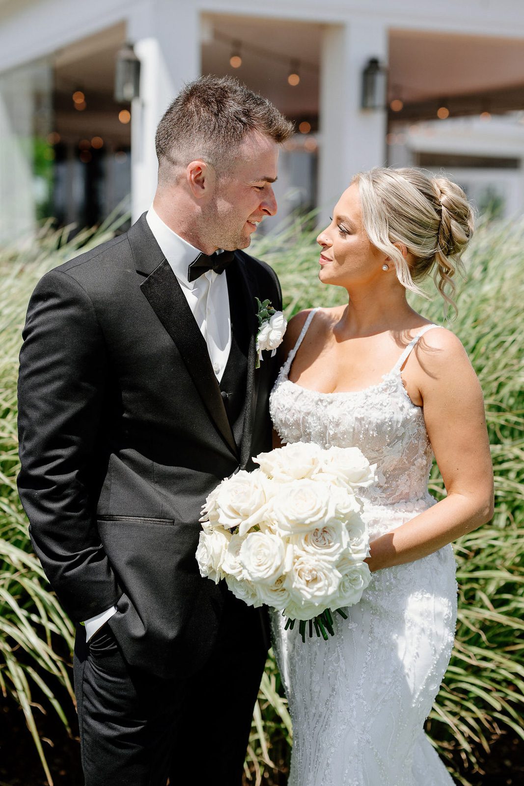 newlyweds smiling in front of a bush and an outdoor seating venue with string lights. 